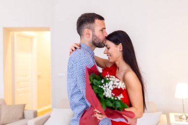 Sevgililer Günü 'nde romantik bir randevuda, arka planda sıcak lamba ışığı olan, samimi, modern bir dairede sevgilisinin alnından bir buket kırmızı gülle öpen bir adam.