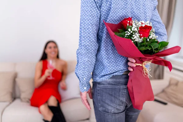 Arkasında bir buket kırmızı gül tutan bir adam kırmızı elbiseli kız arkadaşına sürpriz yapmak için kanepede romantik bir Sevgililer Günü kutlaması için bekliyor.