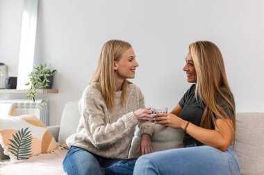 İki mutlu bayan arkadaş evdeki kanepede kahve içip sohbet ediyorlar. Genç kadınlar bir sohbeti paylaşıyor ve parlak bir oturma odasında gülüyorlar. Arkadaşlık kavramı, bağlantı ve rahat yaşam tarzı.