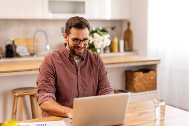 Ahşap bir masada dizüstü bilgisayarıyla uzaktan çalışan neşeli bir adam. Sıcak ışıklandırma, kitap rafları ve sağlıklı ve üretken bir yaşam tarzını yansıtan bir kase meyve ile rahat bir ev..