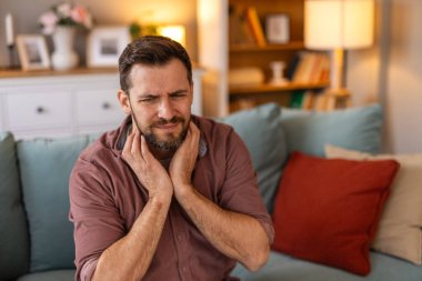 Boynu ağrıyan ya da boğazı ağrıyan kanepede oturan bir erkek. Acı dolu bir ifadeyle boynunu tutan bir yetişkin. Kendini kötü hissediyor, hasta hissediyor ya da evde grip veya soğuk algınlığı belirtileri gösteriyor..