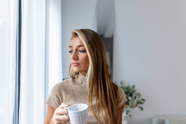 Düşünceli genç bir kadın, parlak bir pencerenin yanında elinde bir fincan kahveyle durup düşünceli bir ifadeyle başka yere bakıyor. Minimalist bir evde yumuşak doğal ışık.
