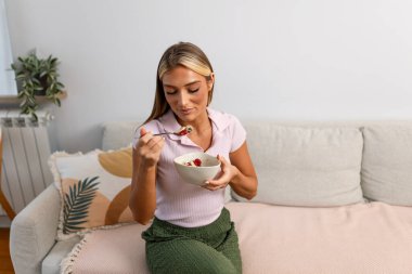 Mutlu bir kadın evdeki kanepede dinlenirken gevrek ve meyveli sağlıklı bir kahvaltı kâsesinin tadını çıkarıyor. Sağlık kavramı, özenli sabah beslenmesi ve modern bir iç mekanda rahat bir yaşam tarzı..