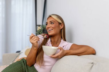 Sağlıklı bir kahvaltı kasesi yerken gülen mutlu genç bir kadın. Olumlu düşünce anlayışı, neşe, sabah refahı ve otantik sağlıklı yaşam, parlak modern ev ortamında..
