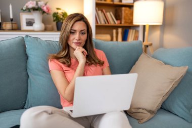 Rahat modern oturma odasında koltukta otururken dizüstü bilgisayar kullanan düşünceli genç bir kadın, evden ya da internetten çalışan bir kadın, uzaktan çalışma ve dijital yaşam tarzı.