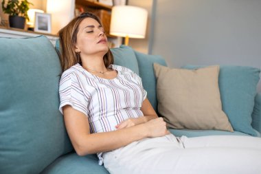 Genç bir kadın kanepeye uzanmış, gözleri kapalı ve midesi ağrılı bir şekilde yatıyor. Kadın ciddi regl krampları ya da evde gıda zehirlenmesi, sağlık sorunları ve fiziksel yorgunluk yaşıyor.