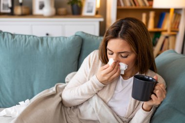 Soğuk ya da mevsimsel alerjiden muzdarip genç bir kadın elinde bir fincan sıcak çay, elinde kağıt mendille burnunu silerken grip tedavisi ve grip tedavisi için battaniyenin altında oturuyor.