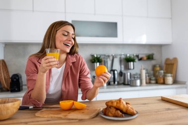 Güzel bir kadın kahvaltıda bir bardak taze portakal suyu içip elinde portakal tutuyor. Sağlıklı sabah alışkanlıkları, doğal vitaminler ve mutlu bir ev hayatı kavramı..