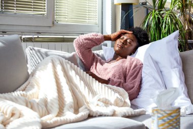 Ateşli, sıcak bir battaniyenin altında kanepede yatan hasta, genç Afrikalı Amerikalı kadın. Evdeki parlak oturma odasında baş ağrısı, soğuk algınlığı ya da grip belirtileri gösteren bir kadın..