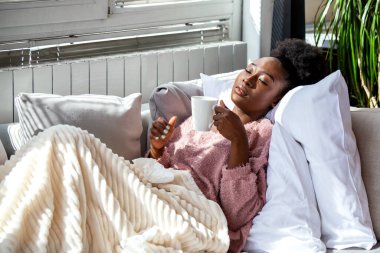 Hasta Afrikalı Amerikalı kadın sıcak bir fincan çayla kanepede dinleniyor. Soğuk algınlığı ya da gripten muzdarip bir kadın, güneşli bir oturma odasında mendillerle battaniyeye sarılmış..