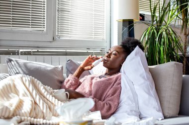 Hasta Afro-Amerikalı kadın kanepede uzanıyor ve burun spreyi ile tıkanıklığı gideriyor. Genç bir kadın soğuk algınlığı ya da grip belirtileri yaşıyor. Parlak bir oturma odasında..