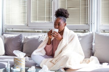 Genç Afro-Amerikalı kadın sıcak bir battaniyeye sarılıp kanepede oturmuş sıcak çay içiyor. Hasta kadın soğuk algınlığı veya gripten kurtulurken evde dokular ve ilaçlarla dinleniyor..