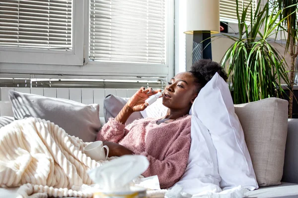 Hasta Afro-Amerikalı kadın kanepede uzanıyor ve burun spreyi ile tıkanıklığı gideriyor. Genç bir kadın soğuk algınlığı ya da grip belirtileri yaşıyor. Parlak bir oturma odasında..