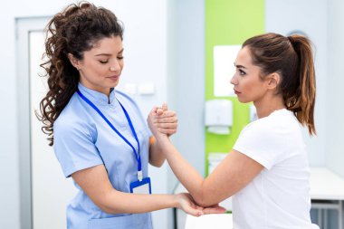 Mavi üniformalı kadın terapist, parlak bir tıbbi tesiste, genç bir kadının kolu ve dirseği üzerinde fiziksel güç testi ve eklem hareketlilik değerlendirmesi yapıyor..