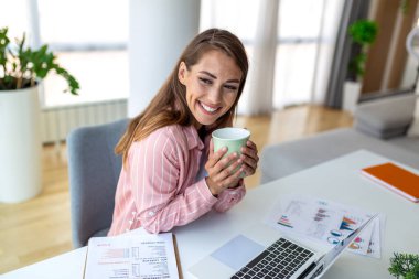 Ev bilgisayarında çalışırken kahve içen mutlu bir iş kadını. Gülümseyen bayan girişimci uzaktan çalışma sırasında sabah çayının keyfini çıkarıyor. Masada güzel bir kız var. Belgeleri ve bilgisayar teknisyeniyle.