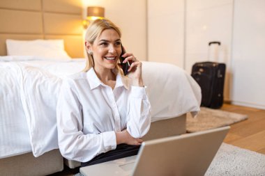 Otel odasında çalışırken telefonda konuşan başarılı bir profesyonel kadın. İş Gezgini Laptop ve Smartphone ile Yatağa Yakın, Uzaktan Çalışma ve Modern İçişlerinde Şirket Seyahati Konsepti Kullanıyor.
