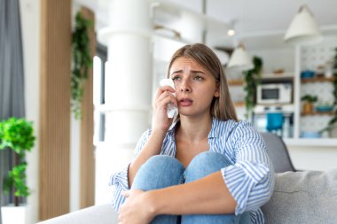 Üzgün kadın kanepede ağlıyor. Akıl sağlığı ve depresyon kavramı. Çekici genç bir bayan kanepeye oturmuş elinde mendille gözyaşlarını siliyor. Evde duygusal stres, keder ve kalp kırıklığı.