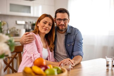 Mutfakta oturup kameraya bakan mutlu çiftin portresi. Genç adam ve kadın evlerinde meyvelerle kucaklaşıyorlar. Modern evde aile hayatı, ilişki ve aile bağları kavramı.