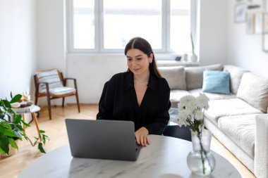 Evde dizüstü bilgisayarda çalışan esmer bir kadın. Modern Parlak Oturma Odası 'nda dizüstü bilgisayar kullanarak Kara Gömlekli Profesyonel Kadın Serbest Çalışan. Uzak Çalışma ve Kariyer Konsepti.