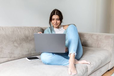 Aydınlık bir dairede koltukta otururken dizüstü bilgisayar kullanan genç bir kadın. Mavi hırkalı bir bayan öğrenci çevrimiçi çalışıyor ya da uzaktan bilgisayar başında çalışıyor. Modern ev ofisi ve eğitim.