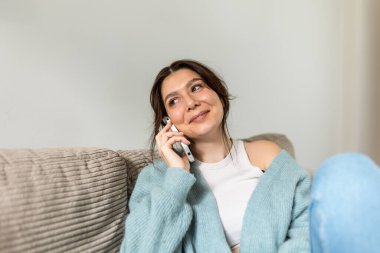 Evdeki koltukta oturmuş akıllı telefondan konuşan gülümseyen genç bir kadın. Mavi hırkalı mutlu kadın serbest yazar, parlak oturma odasında hoş bir telefon konuşması yapıyor. Modern yaşam tarzı ve iletişim