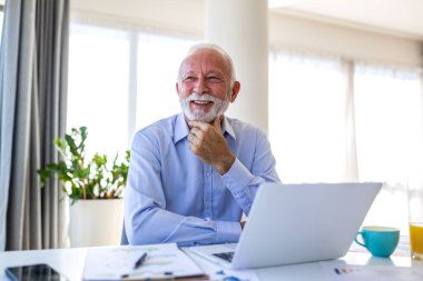 Ofis masasında dizüstü bilgisayarla oturan mutlu kıdemli işadamı. Başarılı yaşlı erkek yönetici gülümseyip iş hedeflerini düşünüyor. Parlak ev ofisinde kendinden emin bir emekli profesyonel..