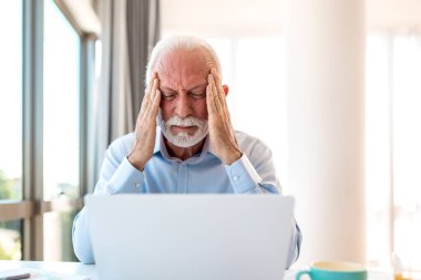 Dizüstü bilgisayarda baş ağrısı olan stresli kıdemli iş adamı. Yaşlı erkek yöneticinin şakaklara dokunması ve ofis masasında migren ağrıları çekmesi. Çok çalışan profesyonel ve tükenmiş..