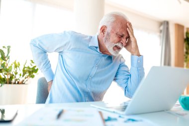 Ofiste sırt ağrısı ve baş ağrısı olan kıdemli bir işadamı. Yaşlı bir yönetici dizüstü bilgisayar kullanırken belini ve alnını acı içinde tutuyor. Kronik ağrı, omurilik problemi ve stres konsepti.