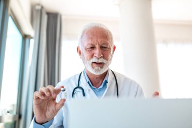 Dizüstü bilgisayardaki tele-sağlık video görüşmesi sırasında elinde aşı şişesi tutan kıdemli bir erkek doktor. Profesyonel yaşlı hekim, hastaya çevrimiçi danışma yoluyla ilaç veya klinik deney sonuçlarını açıklıyor.