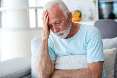 Baş ağrısı ve migren. Alnında ellerle oturan üzgün kıdemli adam, stresi ve akıl sağlığı sorunu olan insanlar, sağlık ve tıp konsepti. Yorgun erkek, kanepede kendini kötü hissediyor..