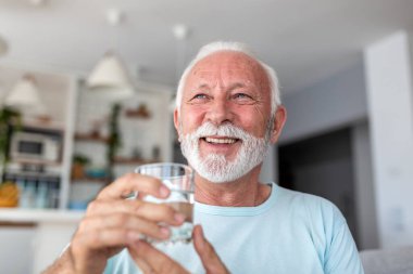 Kıdemli adam su içiyor. Gülümseyen yaşlı erkek elinde bir bardak tatlı su, sağlık sigortası ve ilaç konsepti tutuyor. Mutlu emekli adam evde susuz kalıyor, yaşlılar için sağlıklı bir yaşam tarzı ve sağlık..