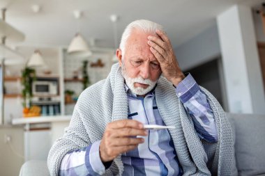 Ateş ve grip belirtileri. Kıdemli adam termometreyle ısıyı ölçüyor ve başını ağrıtıyor. Kanepeye, mevsimlik virüse, sağlık hizmetlerine ve tıp konseptine sarılmış hasta yaşlı erkek..