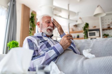 Yüksek ateş ve baş ağrısı. Kanepede termometreyle yatan ve alnında ıslak kompres olan yaşlı bir adam. Evde grip ve mevsimsel virüsten muzdarip yaşlı erkek hasta, sağlık ve tıp konsepti.