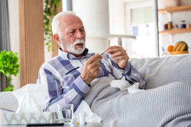 Sıcaklığı kontrol ediyorum. Kanepede yatarken termometreye endişeli bir ifadeyle bakıyor. Hasta yaşlı erkek grip belirtileri, mevsimsel virüs, sağlık ve tıp konsepti evde.