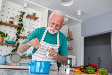 Mutfakta yemek pişirirken çömleğe tuz ekleyen mutlu kıdemli adam. Evde önlük çeşnili yemek yiyen yaşlı bir aşçı. Emeklilik hayatı tarzı. Büyükbaba taze organik sebzelerle sağlıklı bir yemek hazırlıyor..