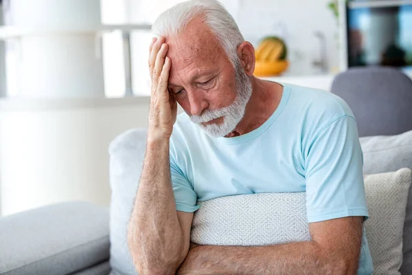 Baş ağrısı ve migren. Alnında ellerle oturan üzgün kıdemli adam, stresi ve akıl sağlığı sorunu olan insanlar, sağlık ve tıp konsepti. Yorgun erkek, kanepede kendini kötü hissediyor..