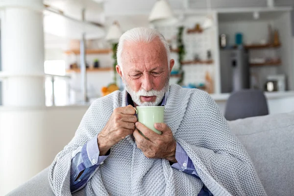 Grip belirtileri gösteren yaşlı bir adam kanepeye oturmuş battaniyeye sarılmış ve elinde bir fincan sıcak çay tutuyor. Hasta, yaşlı bir erkek üşüyormuş, ateşi varmış ya da evde mevsimsel virüs varmış. Sağlık hizmetleri kavramı.
