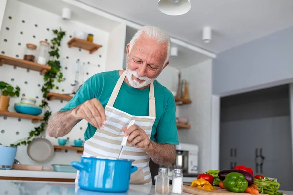 Mutfakta yemek pişirirken çömleğe tuz ekleyen mutlu kıdemli adam. Evde önlük çeşnili yemek yiyen yaşlı bir aşçı. Emeklilik hayatı tarzı. Büyükbaba taze organik sebzelerle sağlıklı bir yemek hazırlıyor..
