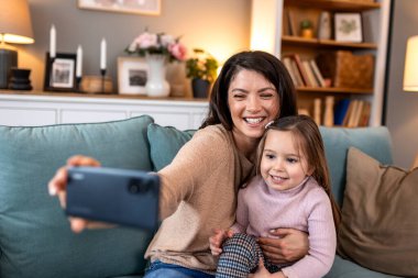 Mutlu anne ve kız akıllı telefonuyla selfie çekiyor. Kanepede fotoğraf için poz veren gülümseyen kadın ve küçük kız. Aile, teknoloji ve rahat bir evde hatıralar yakalama kavramı..