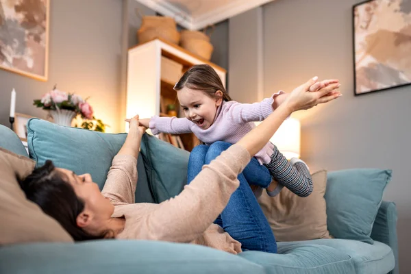 Mutlu anne, küçük kızıyla kanepede uçak oyunu oynuyor. Oturma odasında küçük bir kızı kaldırırken gülümseyen kadın. Aile eğlencesi, kaynaşma ve rahat bir ev içinde eğlenceli yaşam tarzı konsepti.