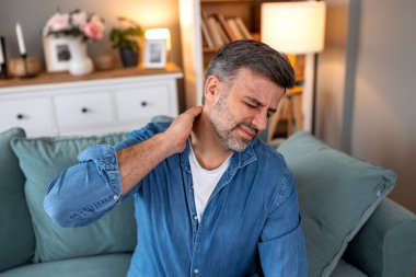 Boyun ağrısı ve kas zorlanması. Yetişkin bir adam kanepede oturuyor ve elleriyle boynunu tutuyor. Sağlık ve tıp konsepti. Yakışıklı erkek yorgun ve rahim ağrısı çekiyor..