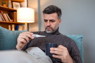 Adam termometreyle ateşi kontrol ediyor. Yetişkin erkek kanepede oturup dijital termometreye bakıyor. Sağlık ve tıp konsepti. Yüksek ateş, grip veya viral enfeksiyon geçiren kişi.