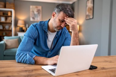 Başı ağrıyan stresli bir iş adamı. Evde dizüstü bilgisayarda çalışan yorgun bir adam, Sağlık ve Tıp konsepti. Olgun erkek serbest çalışan alnına dokunuyor ve iş ya da borç altında eziliyor..