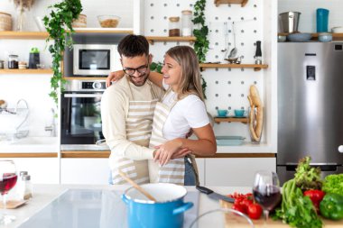Modern mutfakta yemek pişirirken kucaklaşan mutlu genç çift. Önlüklü kadın ve erkek sarılıp gülüyorlar. Romantizm ve yaşam tarzı kavramı. İnsanlar taze sebzelerle yemek hazırlıyorlar..