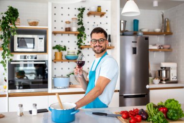 Mutfakta yemek pişirirken kırmızı şarap içen önlüklü mutlu adam. Erkek Alkol ve Evde Sağlıklı Yemek Hazırlamak 'ın tadını çıkarıyor. Sağlık ve Yaşam Tarzı Rahatlama ve Diyet Konsepti.