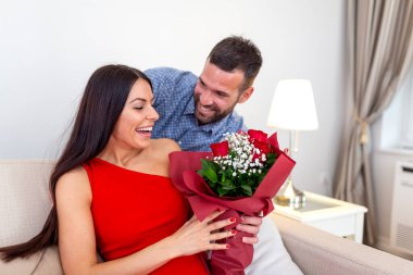 Kırmızı elbiseli güzel bir kadın Sevgililer Günü için bir buket kırmızı gül alıyor. Mutlu genç çift evde çiçekler ve sevgiyle romantik bir randevu gecesini kutluyor..
