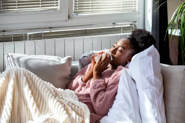 Kanepeye uzanmış, battaniyeli ve sümküren burunlu hasta kadın. Afro-Amerikan Kadın Soğuk algınlığı ya da evde grip. Sağlık ve Tıp Konsepti 'nden. Mevsimsel Alerjiden muzdarip Genç Kadın.