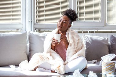 Hasta Kadın, battaniyeye sarılmış, kanepeye çay içiyor, grip ve soğuk sağlık sigortası kavramı. Genç Afrikalı Amerikalı kadın elinde mendil kutusuyla oturma odasında kendini kötü ve yorgun hissediyordu..