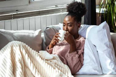 Hasta Afro-Amerikan Kadın Kanepede Çay İçiyor, Soğuk ve Grip Sağlığı Konsepti. Battaniyeye sarılı genç bir kadın elinde kupa, evinde kendini kötü hissediyor. Kucağında mendiller, kış mevsimsel hastalıkları var..