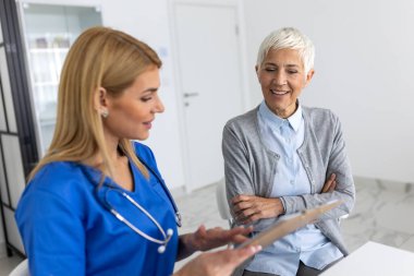 Mavi önlüklü profesyonel kadın doktor, modern bir sağlık kliniğinde özel bir danışmanlık sırasında gülümseyen yaşlı bir kadınla tıbbi sonuçları panoda tartışıyor..